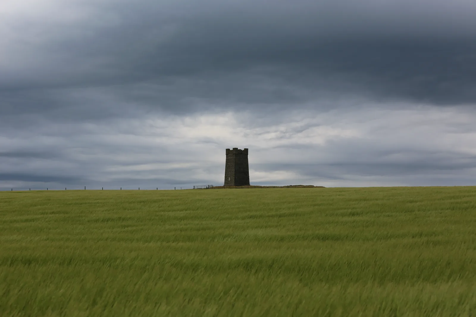 The Kitchener Memorial on Marwick Head seen from inland — a square crenellated stone tower standing alone on the skyline behind a post fence, across a wind-blown field of green barley, under heavy grey cloud