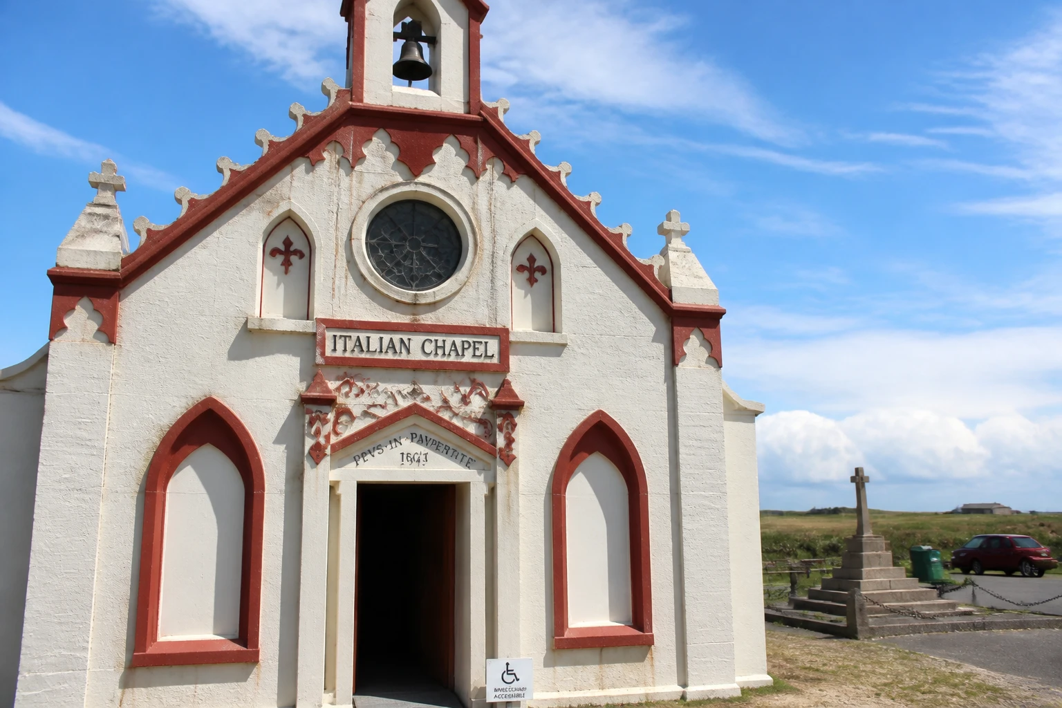 Close-up of the cream and red painted concrete facade of the Italian Chapel on Lamb Holm Orkney, with the three pointed arched entrance, central rose window, and a wheelchair-access sign mounted beside the open doorway, taken on a bright partly-cloudy summer day
