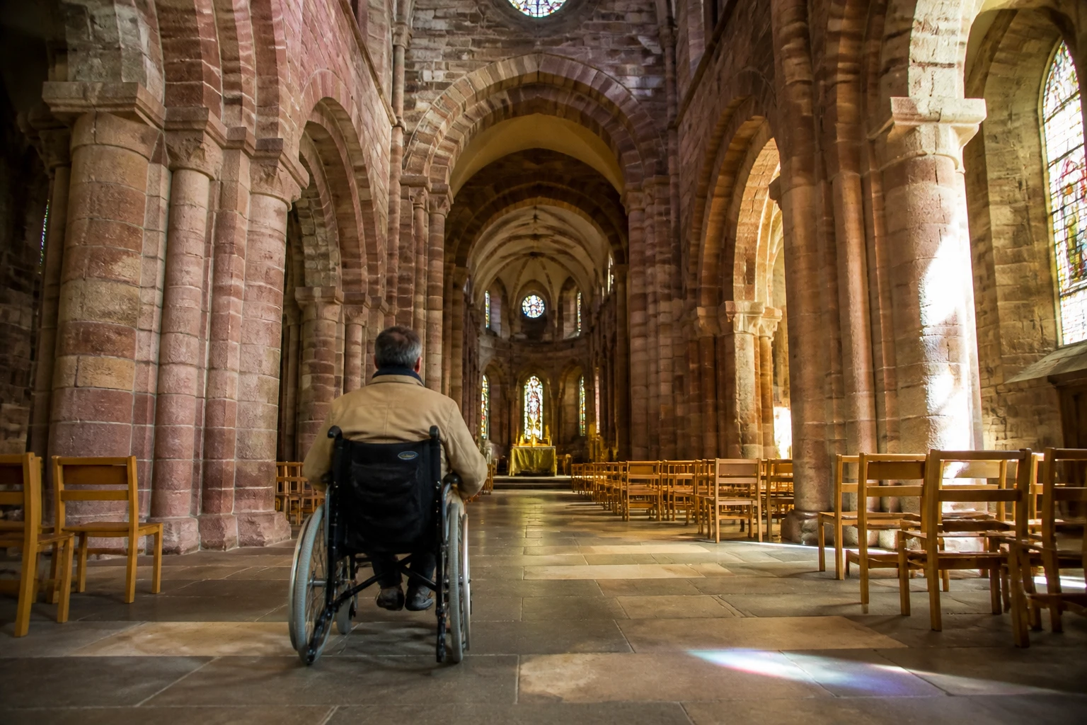 Wheelchair user rolling up the central aisle of St Magnus Cathedral Kirkwall, looking towards the high altar past Romanesque arches and tall sandstone columns, with daylight streaming through high stained-glass windows on the right and rows of wooden chairs on either side of the polished flagstone floor