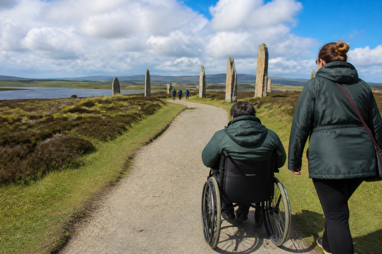 Wheelchair user and a walking companion moving along the firm light gravel path that leads from the visitor car park towards the Ring of Brodgar stone circle on Orkney, with the loch of Stenness on the left, several tall pinkish-grey megaliths in the middle distance and a wide partly-cloudy summer sky overhead