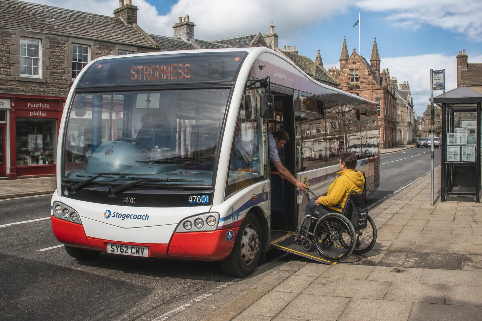 A white-and-red Stagecoach North Scotland single-decker low-floor bus parked at a stop on Broad Street Kirkwall Orkney with the boarding ramp deployed and a wheelchair user in a yellow waterproof jacket about to roll onto the ramp, with the destination display reading STROMNESS visible above the windscreen and red-painted sandstone shop fronts behind