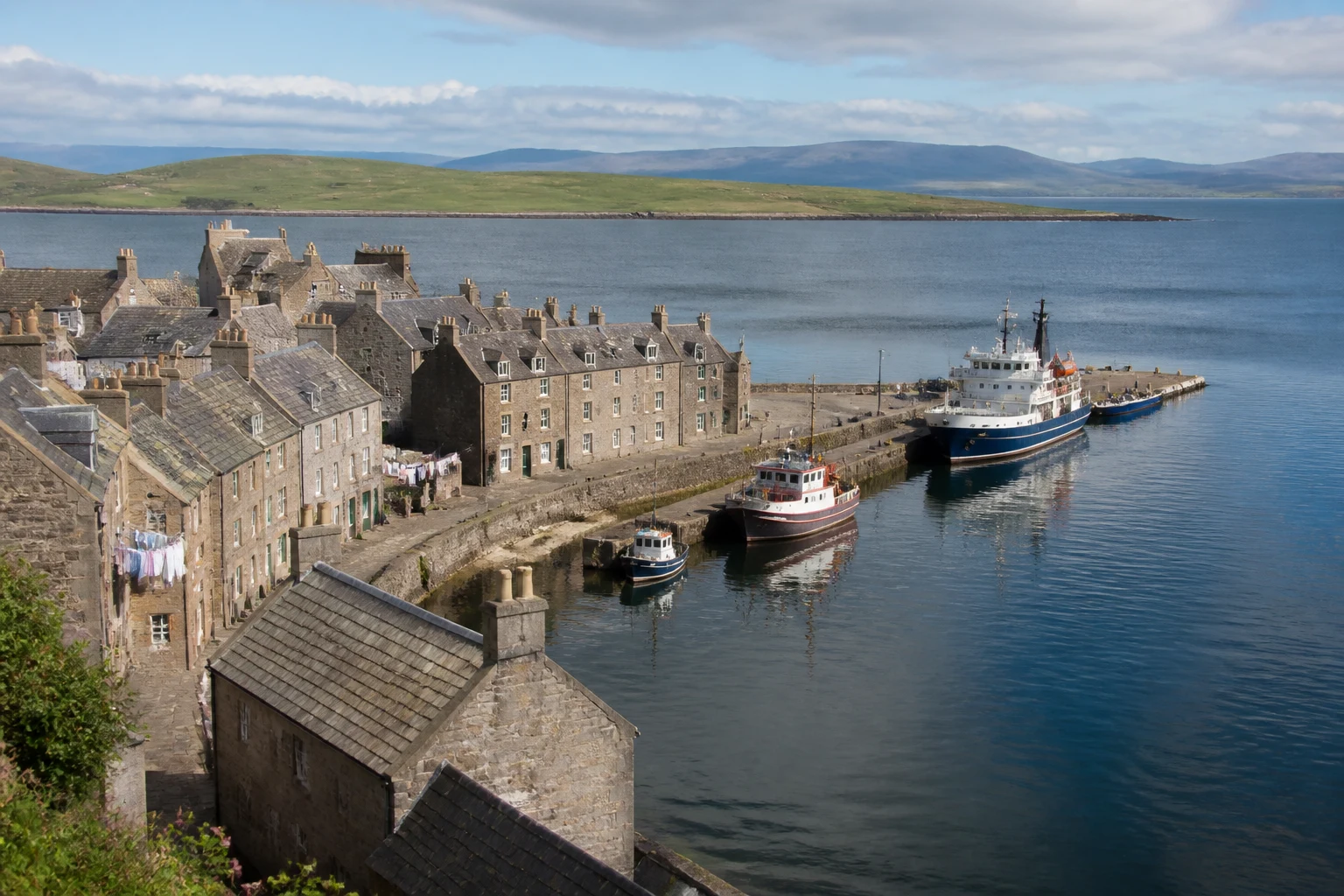 Wide hero view of Stromness harbour Orkney from above with stone flagstone houses curling around the inner harbour water at gold-evening light, the best place to stay in Orkney for atmospheric West Mainland trips