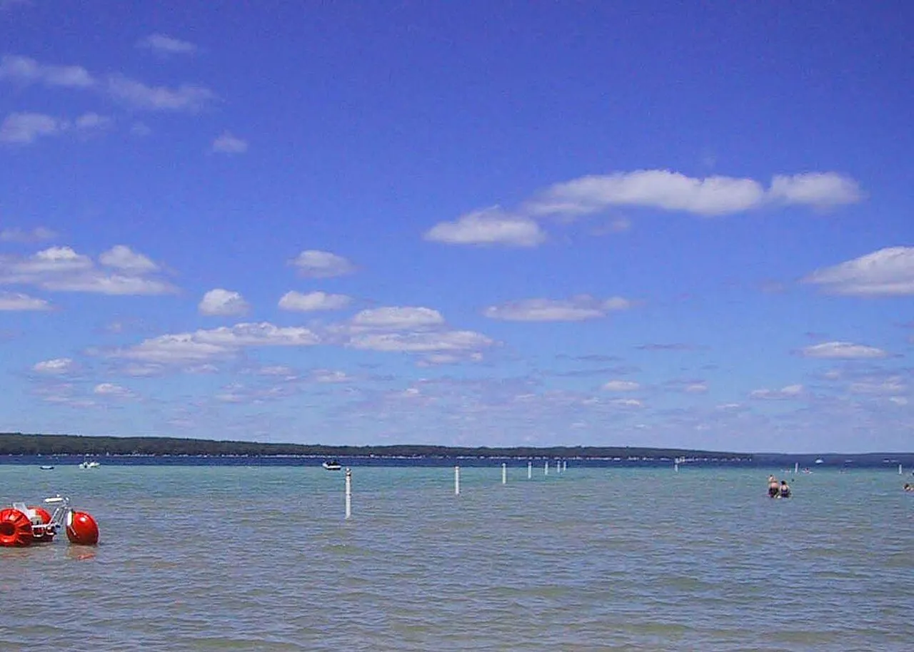 Families swimming in the clear blue water of Higgins Lake, Michigan, with a red paddleboat near the sandy shore