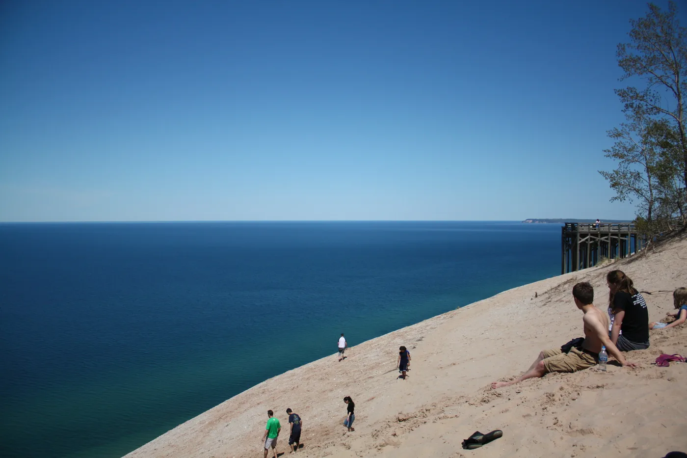 Visitors sitting and walking on the steep sand dunes at Sleeping Bear Dunes National Lakeshore, overlooking a deep blue Lake Michigan under a clear summer sky