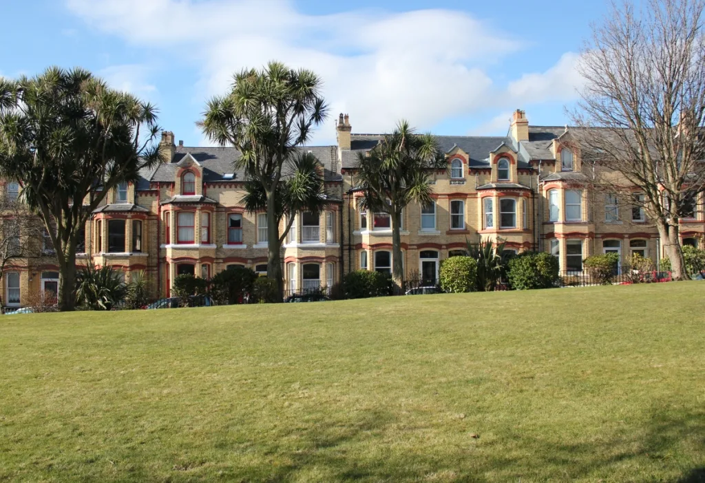 A terrace of tall Victorian houses with bay windows across the lawn of Hutchinson Square in Douglas