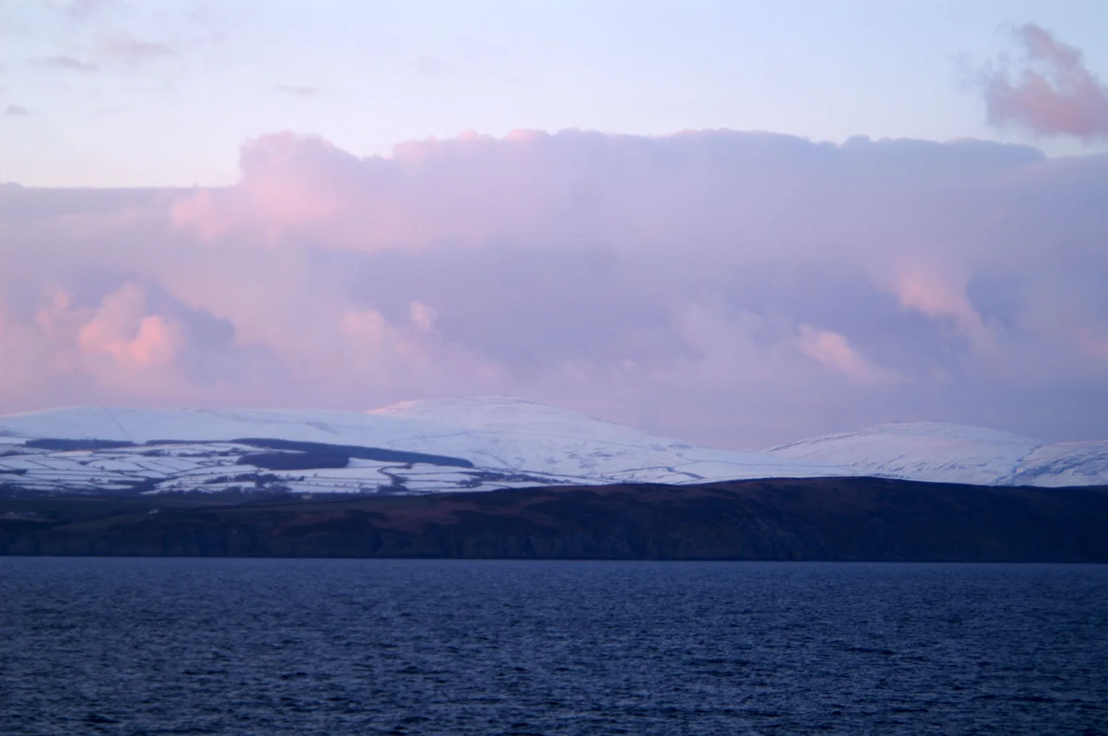 Snow lying on the Manx uplands behind the dark cliffs of Onchan Head, seen from the sea under pink sunset cloud