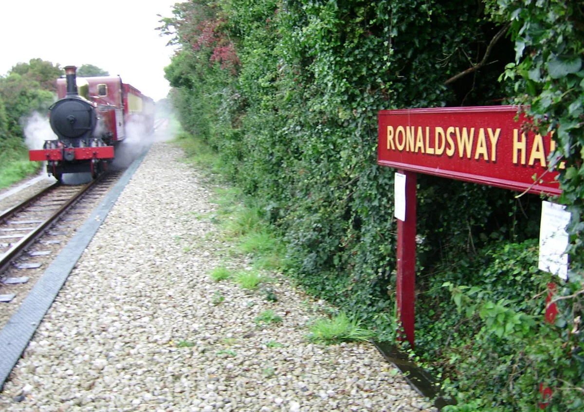 A red and black Isle of Man Railway steam locomotive and carriages approaching the red Ronaldsway Halt sign