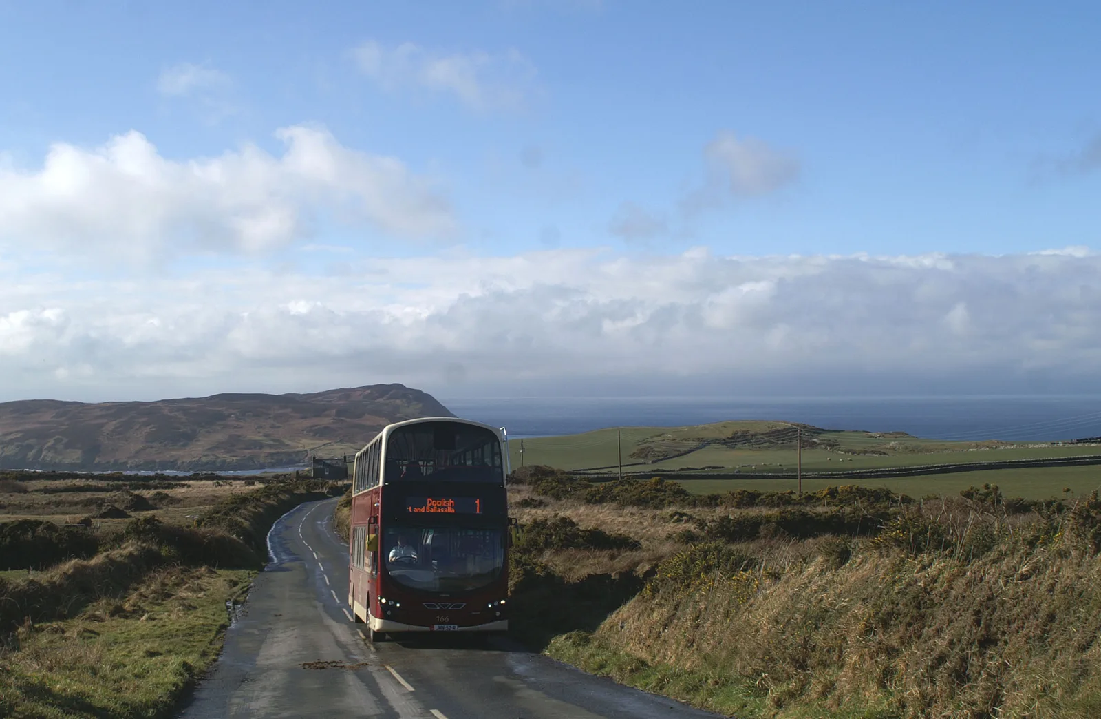 A maroon and cream double-decker bus on a narrow moorland road near Cregneash, with the sea behind