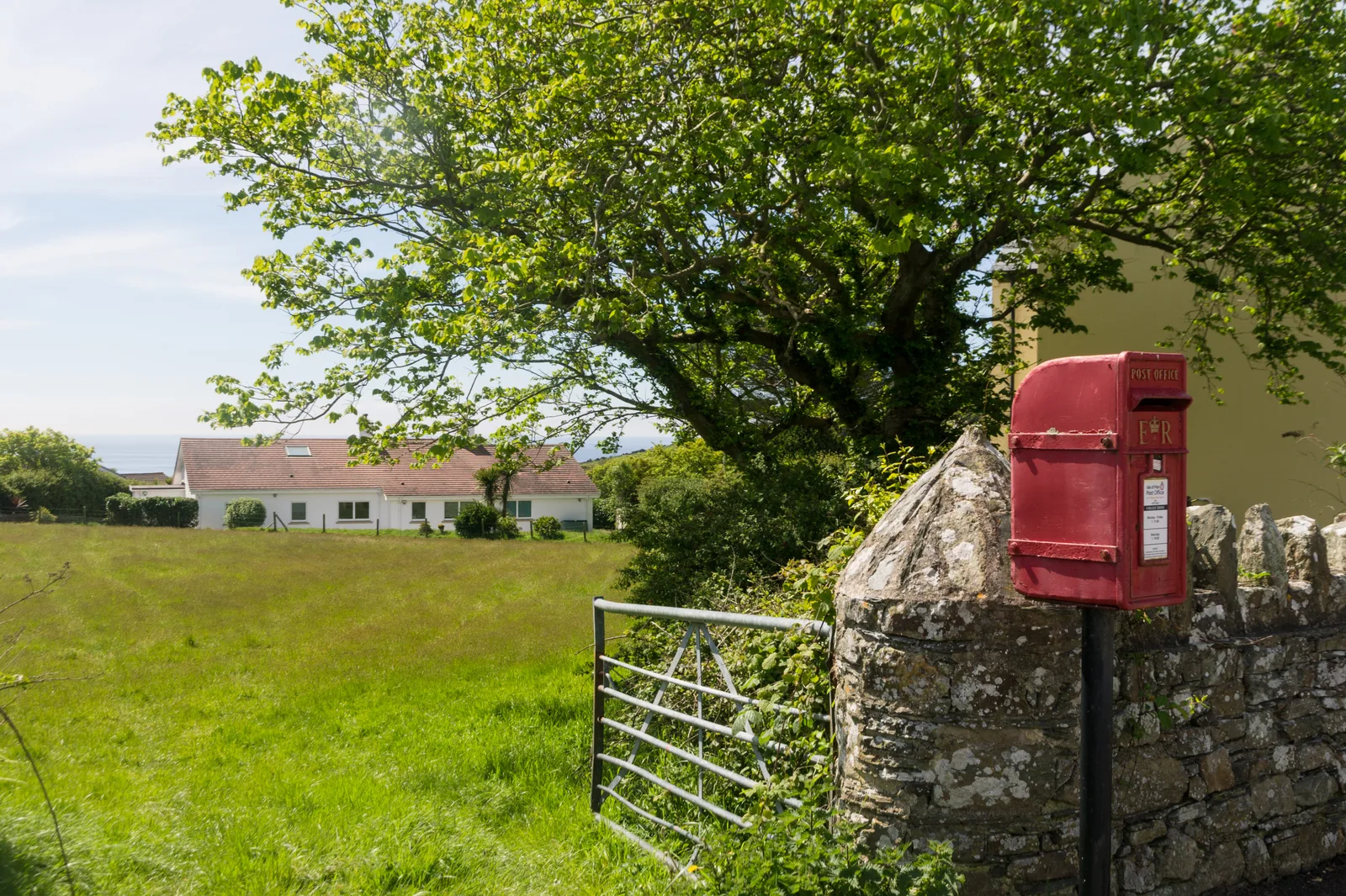 A red wall-mounted postbox on a post beside a stone wall and field gate on a country road