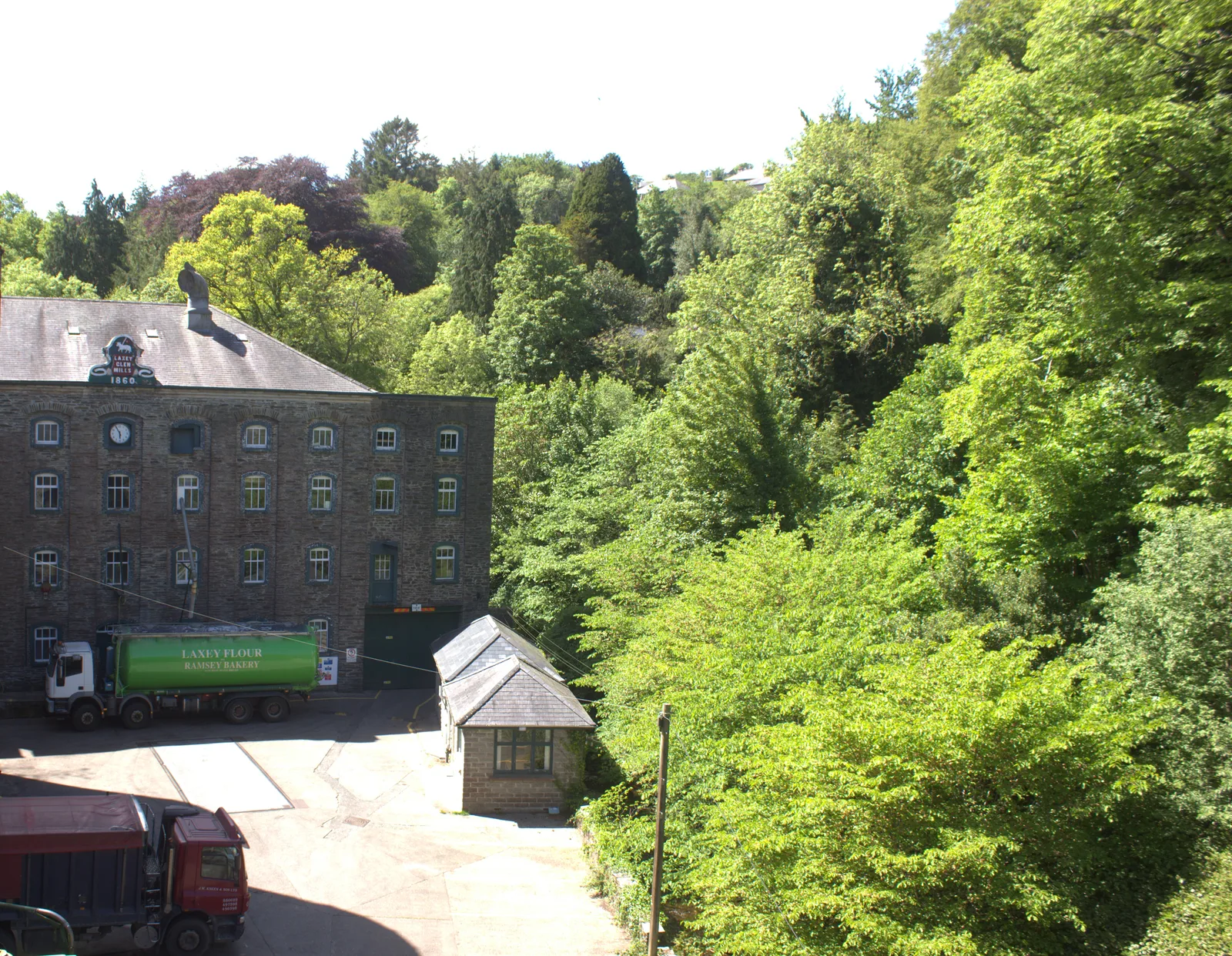 The stone building of Laxey Glen Mills with its 1860 gable sign, a flour tanker in the yard and the wooded glen beside it