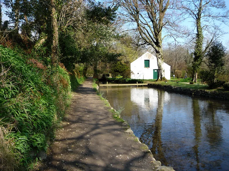 A level path beside still water and trees in Silverdale Glen, with a white stone building beyond