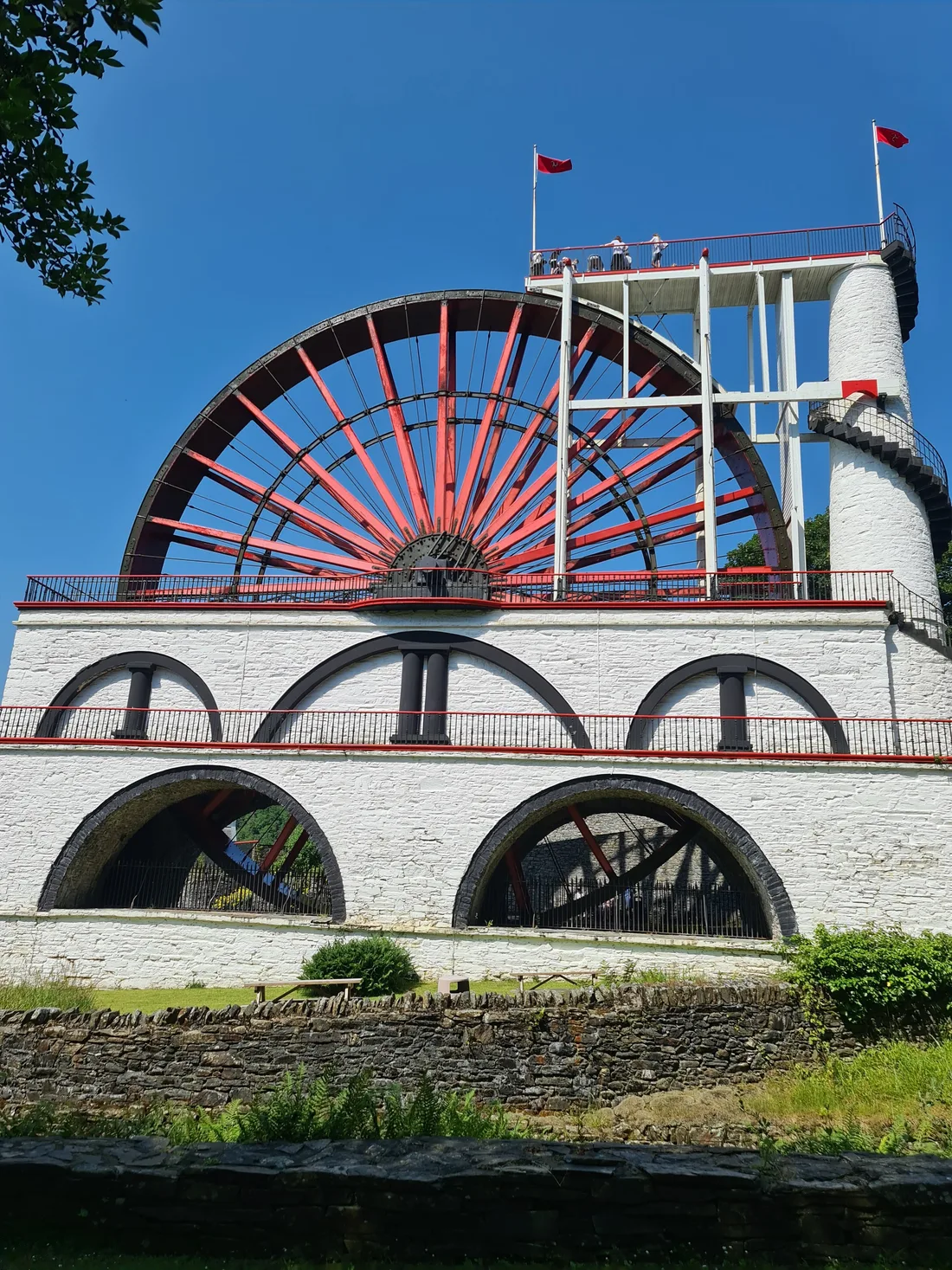 The Great Laxey Wheel (Lady Isabella), its red spokes rising above a whitewashed arched housing, with the spiral staircase tower and viewing platform on the right
