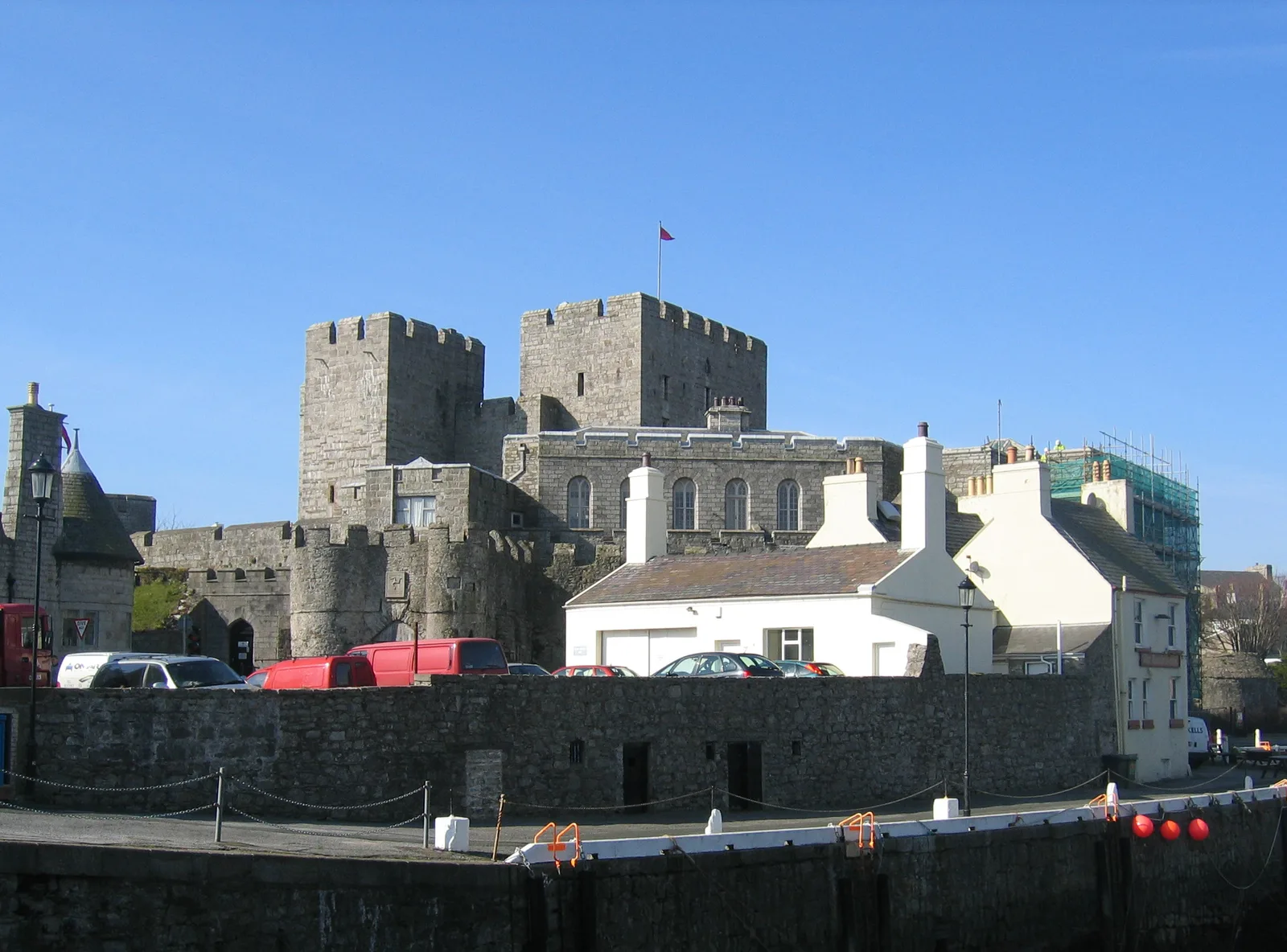 The grey stone towers of Castle Rushen rising behind a white pub and the harbour wall in Castletown