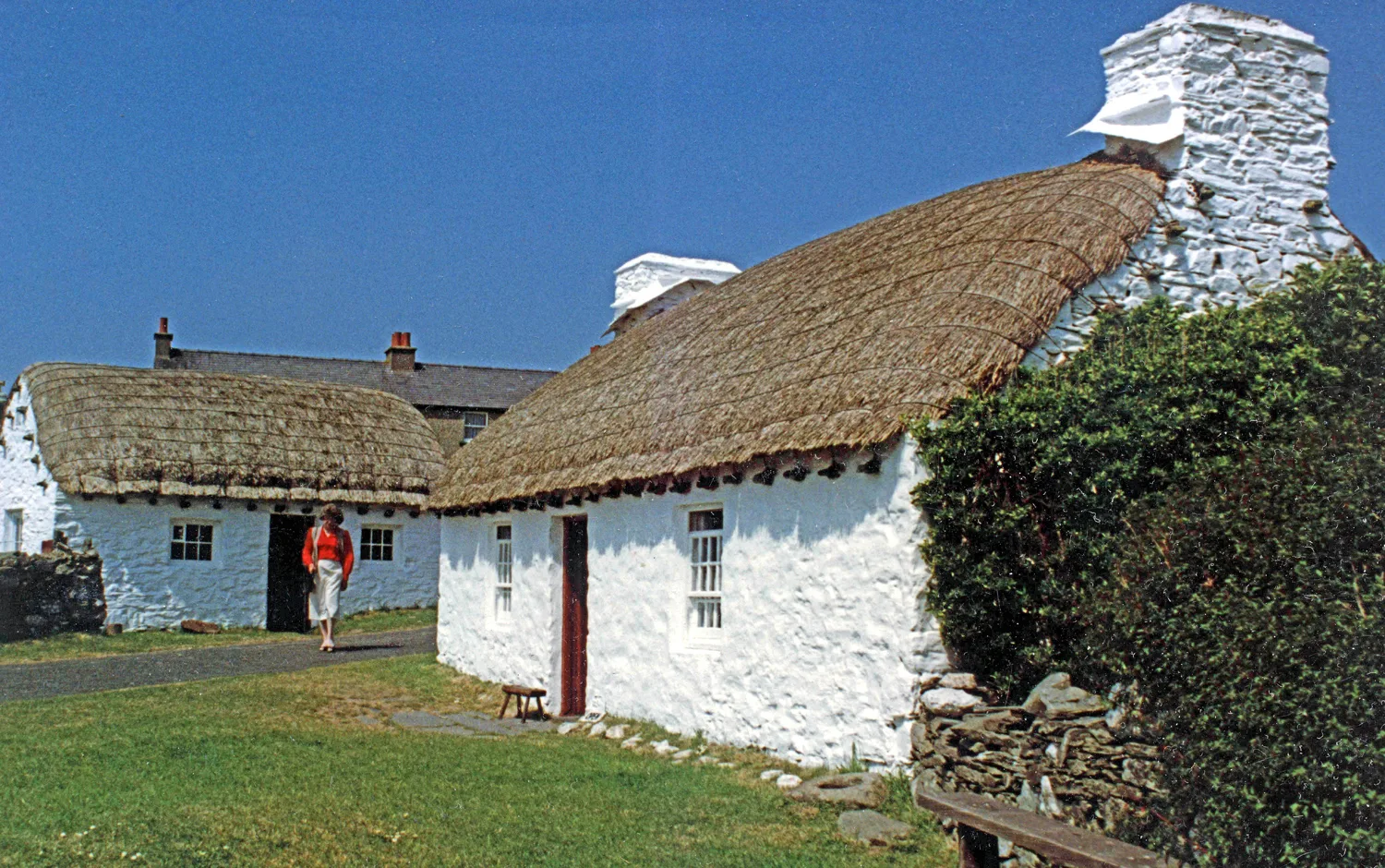 Two whitewashed thatched cottages at Cregneash under a clear blue sky, with a visitor walking between them