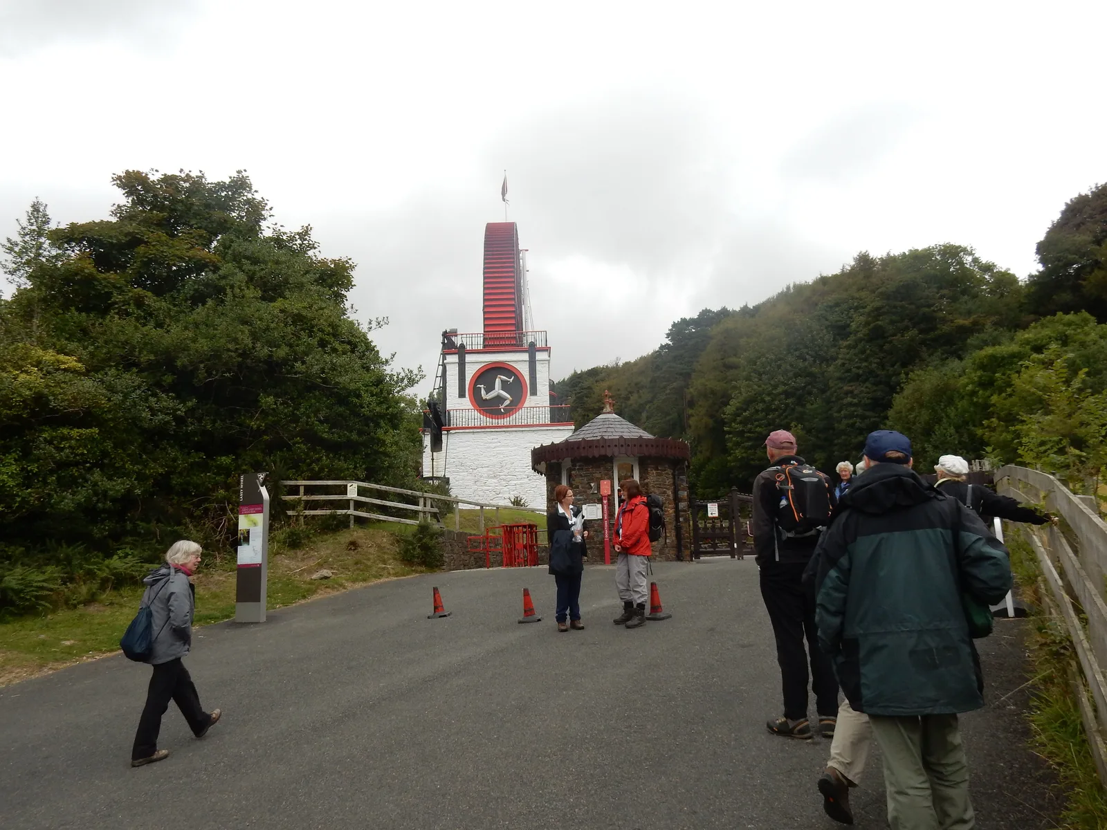 Walkers on the approach road to the Great Laxey Wheel, its red wheel rising above a white tower painted with the Three Legs of Mann