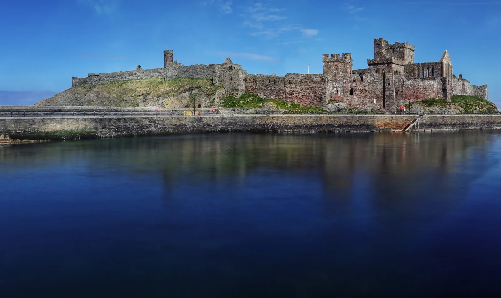 Peel Castle's red sandstone curtain wall and towers on St Patrick's Isle, reflected in the still water of Peel harbour