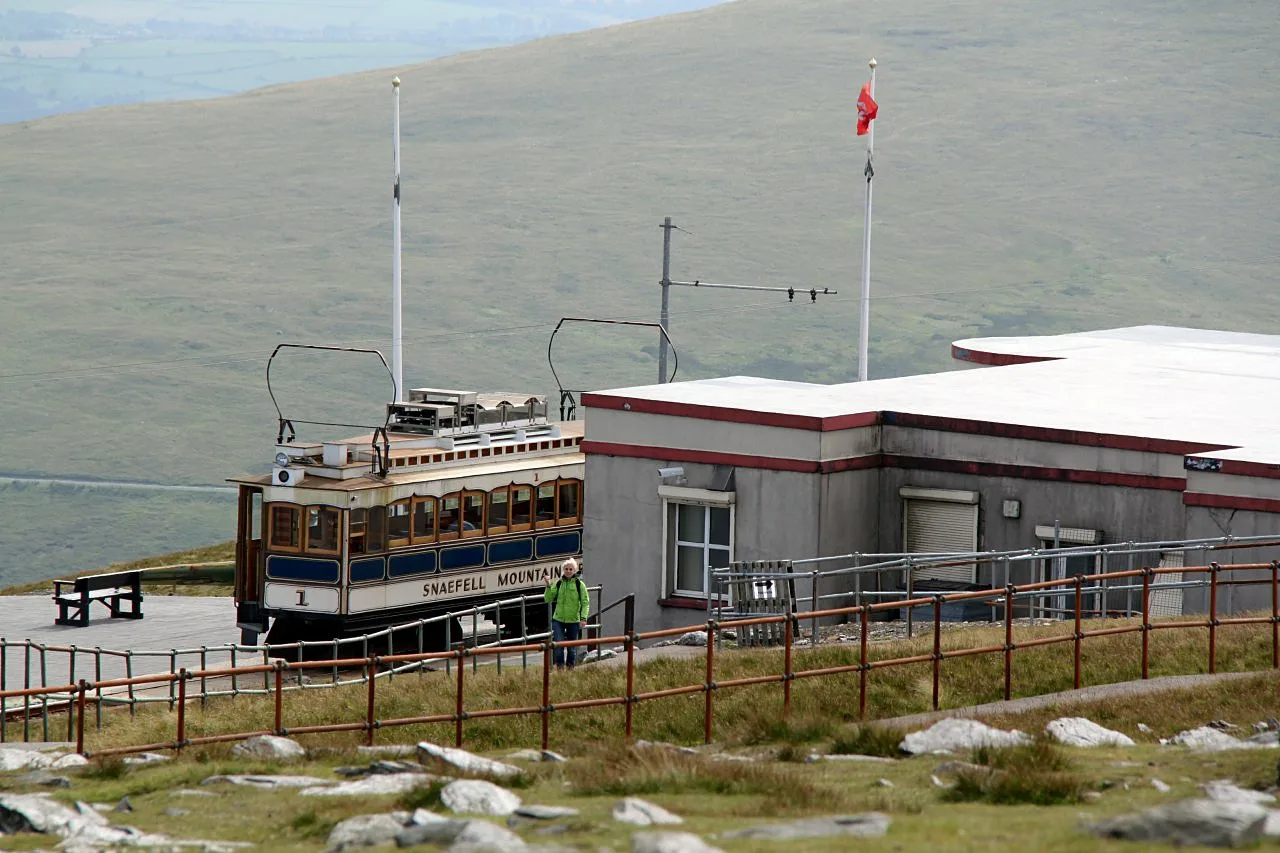 A Snaefell Mountain Railway tramcar standing at the summit station beside the low summit building, with hazy moorland behind