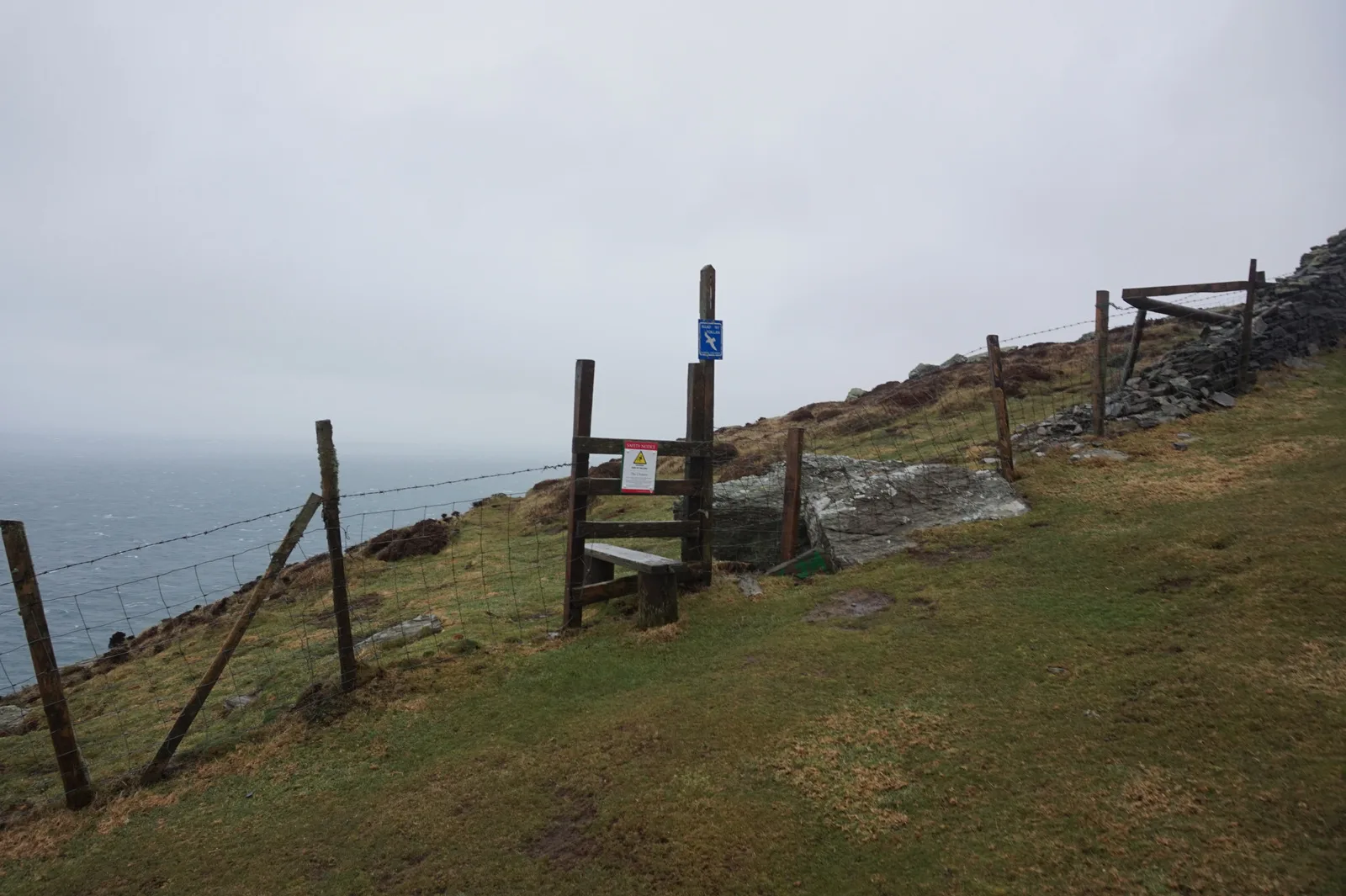 A wooden stile and fence line on a grassy cliff-top section of the coastal path at the Chasms
