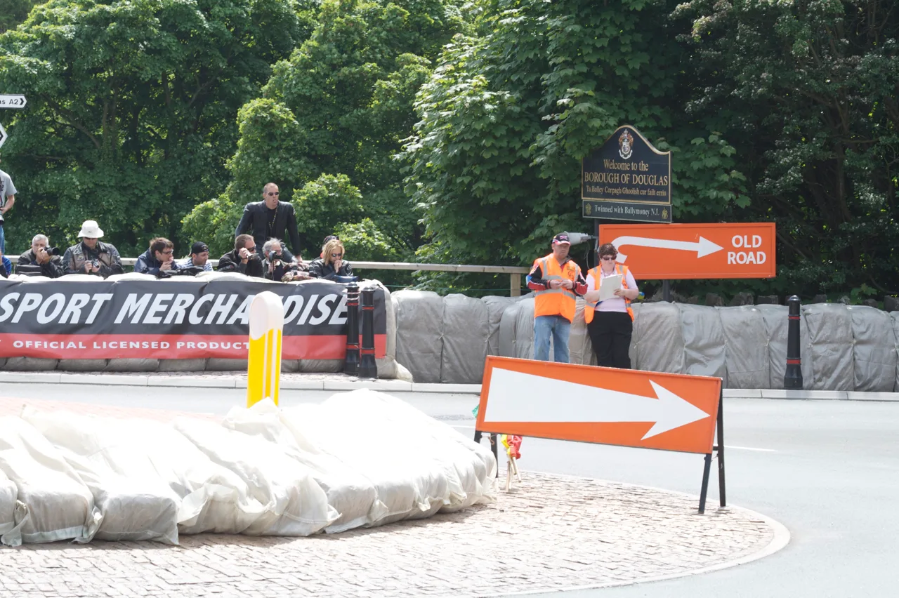 Two marshals in orange tabards, one holding a clipboard, beside orange Old Road arrow signs at Governor's Bridge while spectators with cameras watch from behind a padded barrier