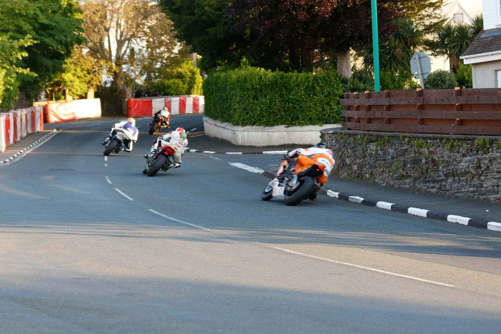 Four racing motorcycles, seen from behind, leaning through a corner on May Hill in Ramsey between hedges and red and white barriers