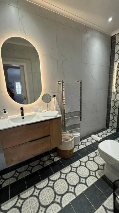 Bathroom with a backlit oval mirror, wood vanity, heated towel rail and patterned tile floor