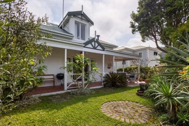 White gabled cottage with wraparound veranda, exposed brick chimney and a lush, established garden
