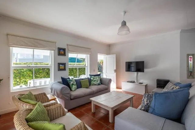 Living room with grey sofas and wicker chairs, looking out through shuttered windows to hedges