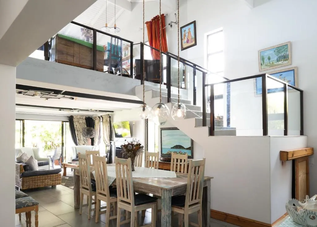 Double-volume dining room with a wooden table for eight beneath an open-tread staircase and mezzanine