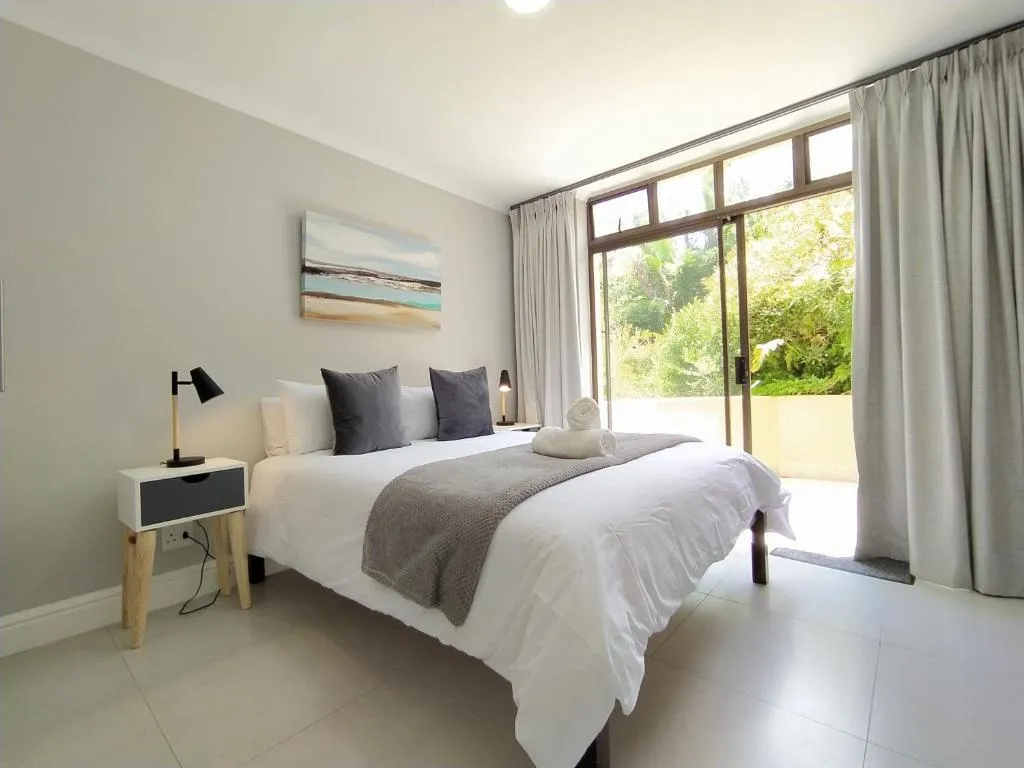 Second bedroom with grey linens, framed beach artwork and a sliding door to a leafy balcony