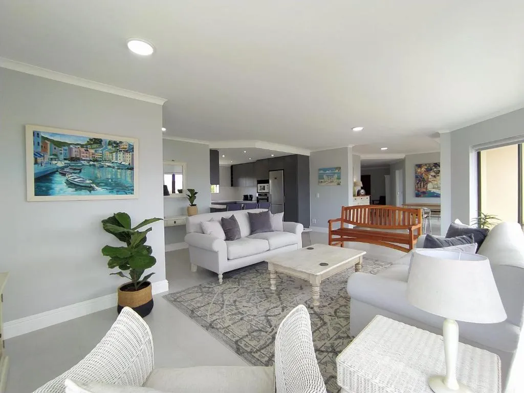 Open-plan living room with white sofas, a harbour-scene painting and access to the kitchen