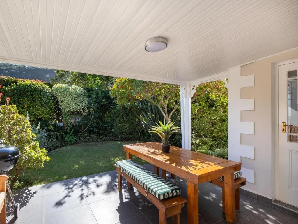 Covered patio with a wooden bench table and braai, backing onto a garden of shrubs and trees