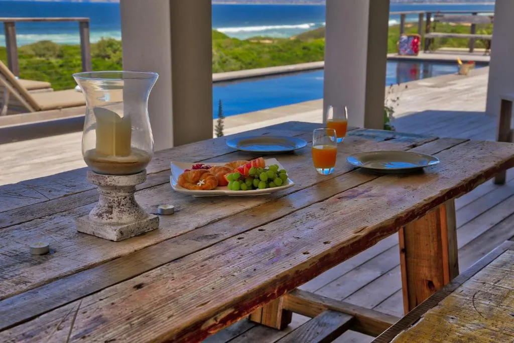 Outdoor dining table set with breakfast, pool and sea visible beyond the pillars