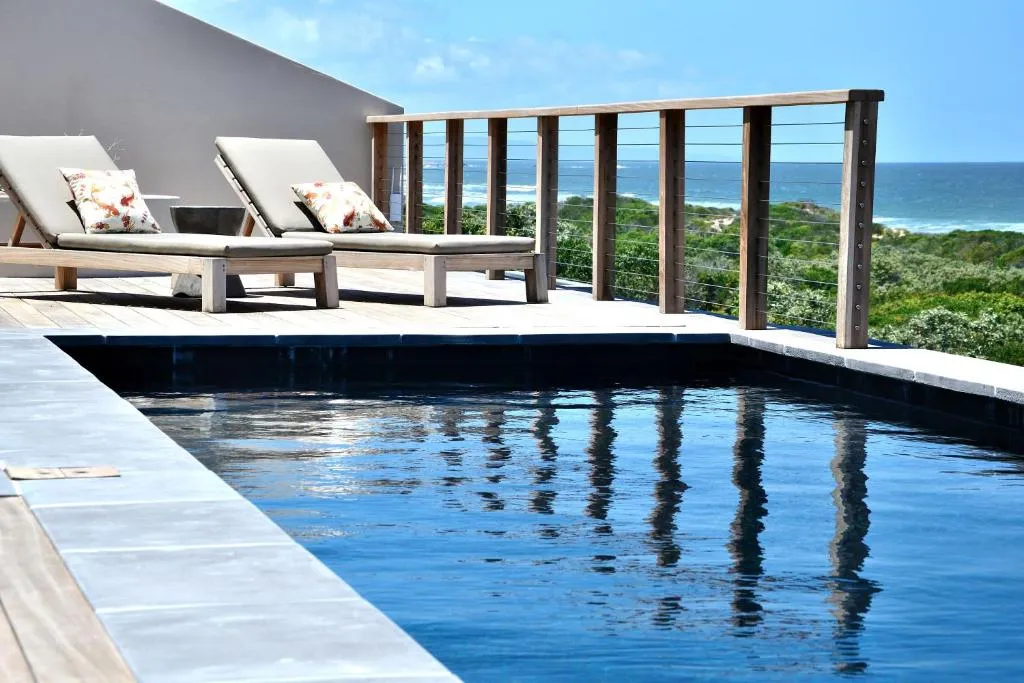 Rooftop pool deck with sun loungers and glass balustrade overlooking the sea beyond dune vegetation