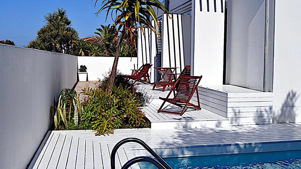 Private pool deck with timber decking, red steamer chairs and tropical planting beside the white house
