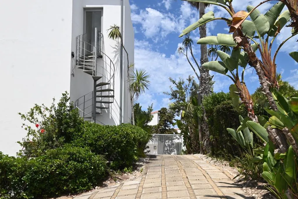 White modern building with a spiral staircase, palms and a paved path leading to the gated entrance