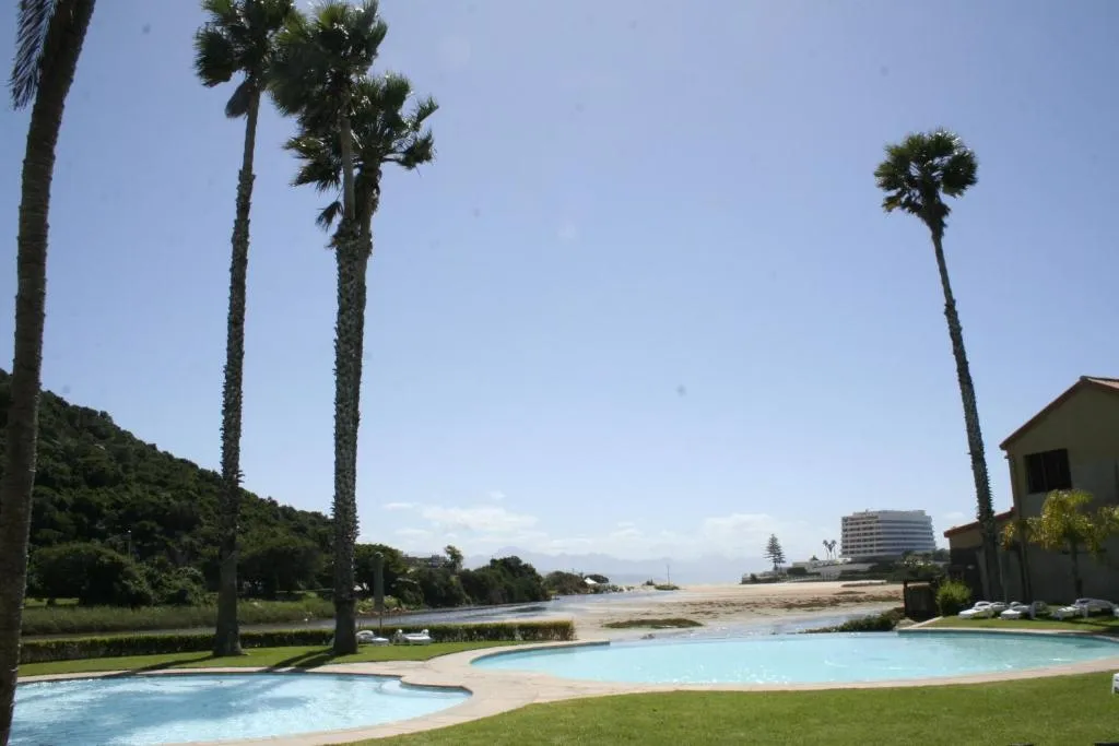 Complex's shared pool and lawn with palm trees, framed by the Piesang River mouth and beach beyond