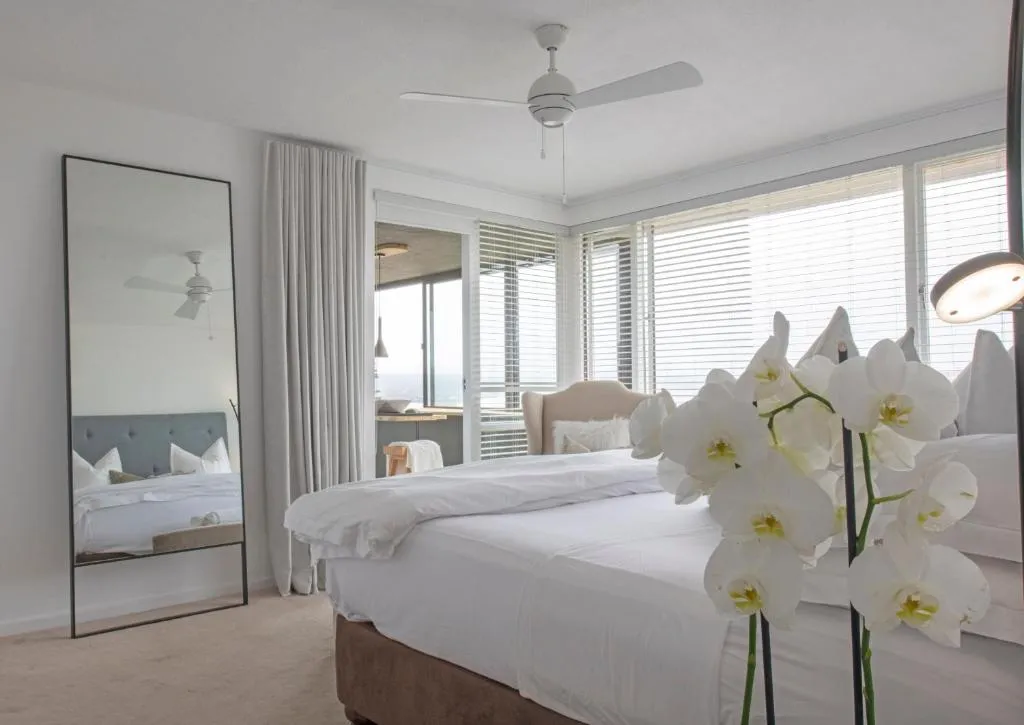 Bright bedroom with a large floor mirror, ceiling fan and louvred windows toward the sea
