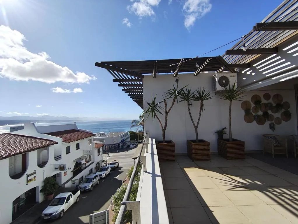 Balcony with potted palms overlooking coastal rooftops, the street below and sea in the distance