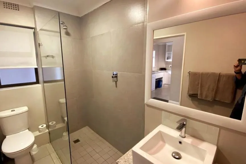 Walk-in shower bathroom with a toilet, sink and a mirror reflecting through to the kitchen