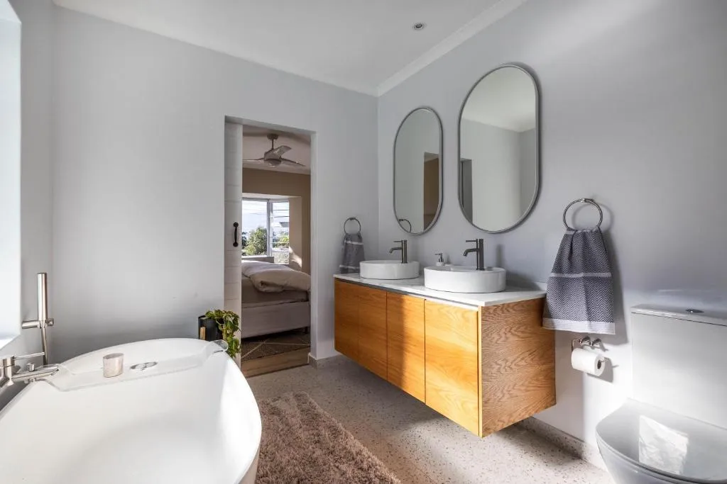 Bathroom with a floating wood vanity, oval mirrors and a tub opening onto the bedroom