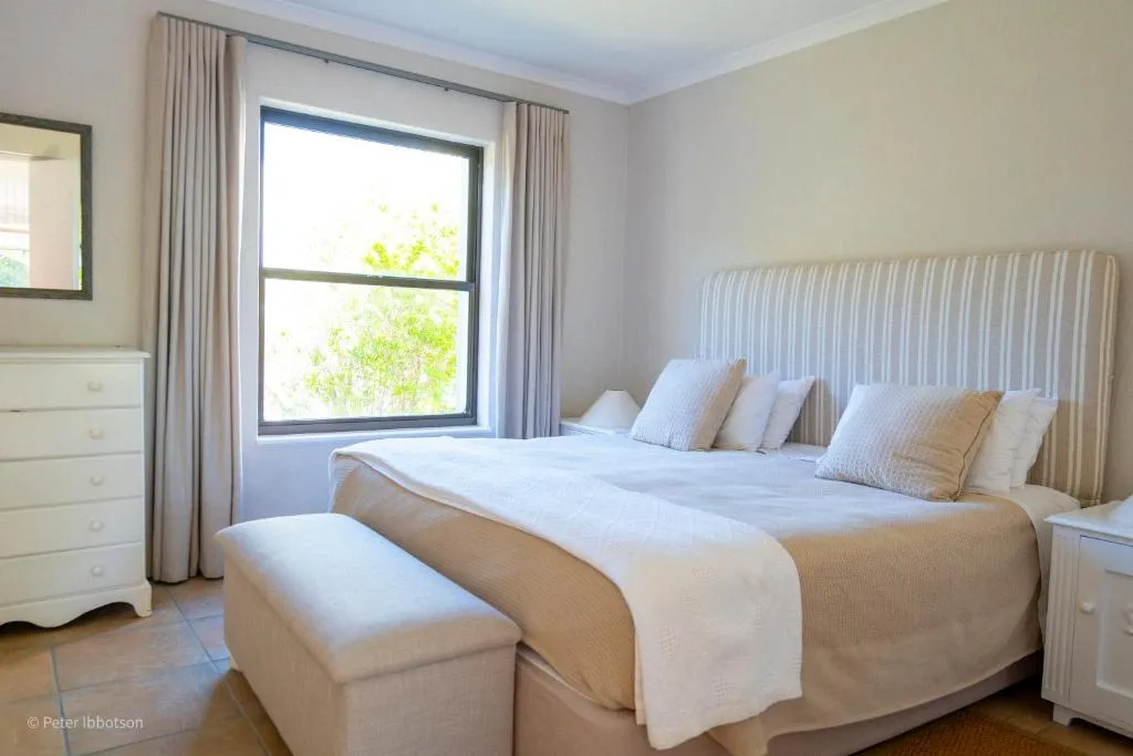 Bedroom with a striped upholstered headboard, white dresser and a window onto leafy trees