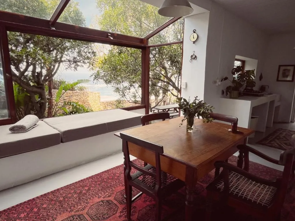 Glass-roofed dining nook with a daybed and sea view framed by trees and a stone garden wall