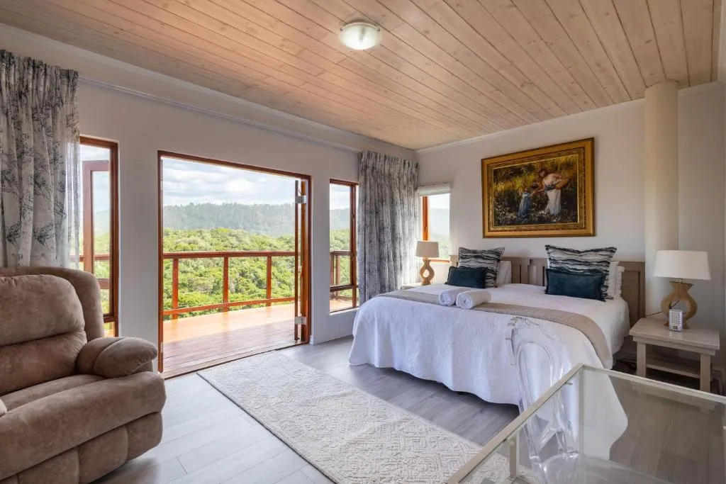 Bedroom opening onto a balcony over a forested valley, with a wingback armchair beside the bed