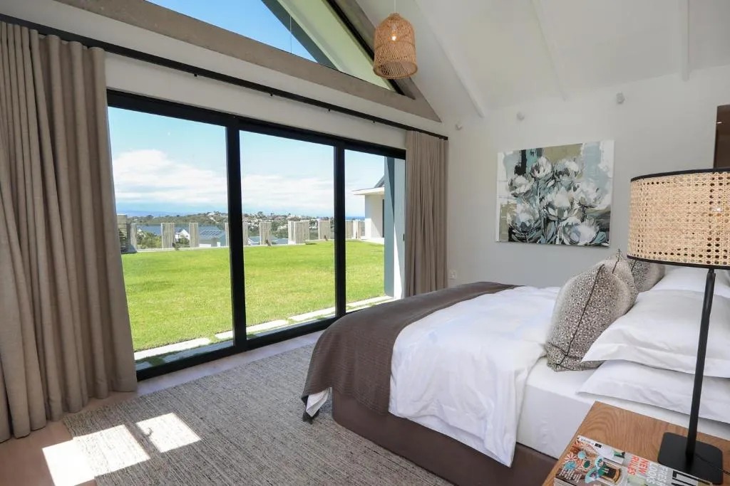Vaulted bedroom with floor-to-ceiling glass doors opening onto a lawn and rooftops with the sea visible beyond