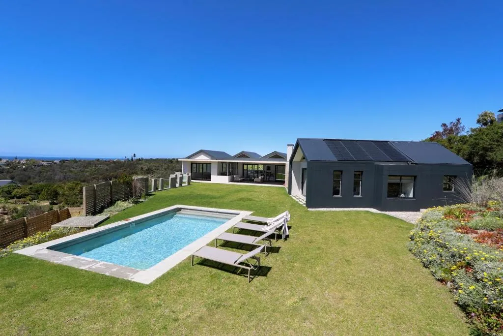 Rectangular pool and lawn beside a black gabled wing of the house, with the coastline visible on the horizon
