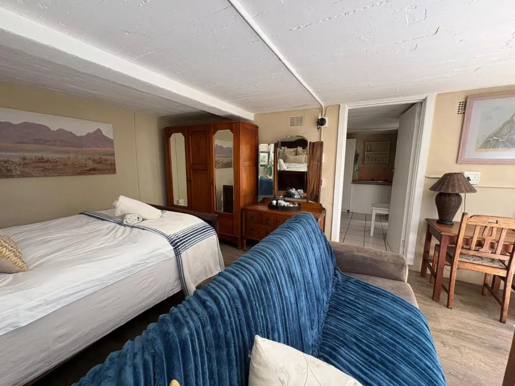 Open-plan studio combining a single bed, blue sofa and dresser with a kitchen doorway beyond