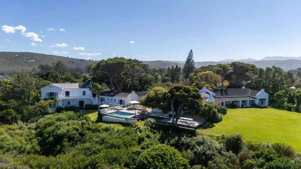 Aerial view of a Cape Dutch-style villa with white gables, a pool and manicured lawn amid green hills