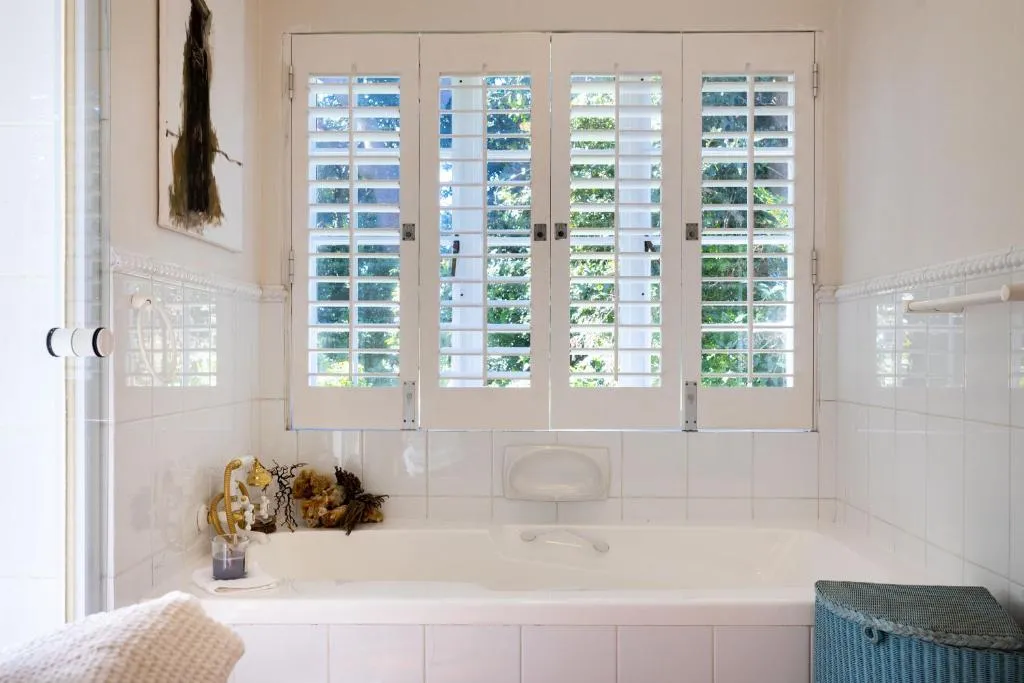 En-suite bathroom with a freestanding bath and louvred plantation shutters framing a leafy outlook