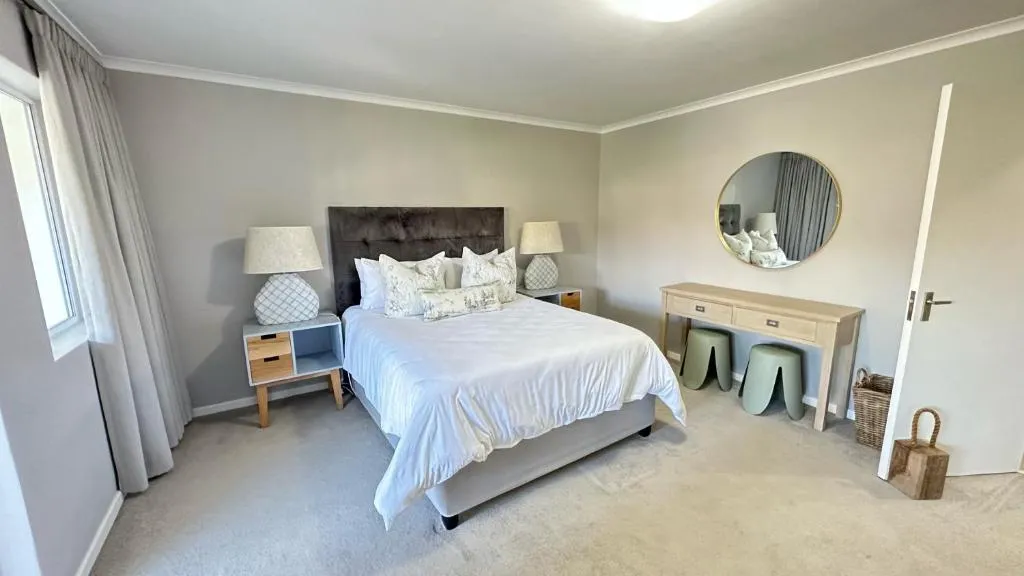 Carpeted main bedroom with a tufted headboard, twin bedside lamps and a round mirror over a console