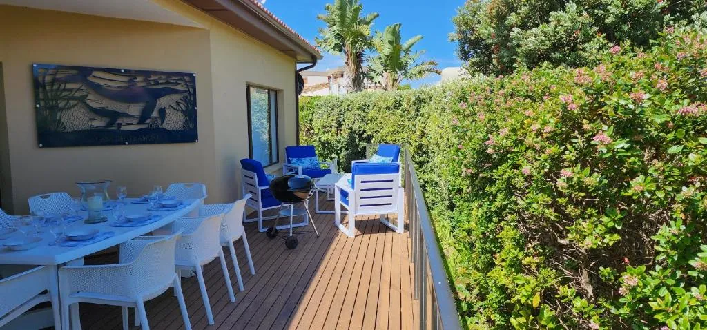 Covered wooden deck with a braai, outdoor dining table, and lounge seating beside a hedge